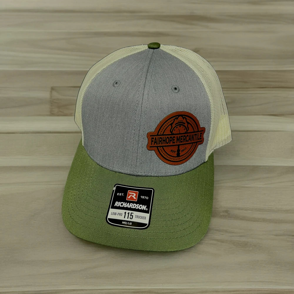 The Fairhope Mercantile Six Panel Patch Hat - Richardson 115 Trucker is a two-tone trucker hat featuring a grey front, green bill, and beige mesh back. It boasts an embroidered patch with "Fairhope Mercantile" on the front and a Richardson 115 label on the bill.