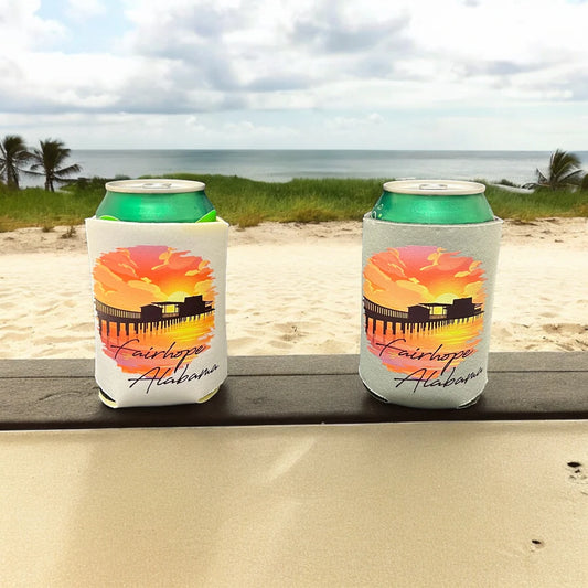 On a wooden ledge overlooking a sandy beach and ocean, two Fairhope Sunset - Can Huggers with "Fairhope, Alabama" sleeves rest under a cloudy sky as the sun sets at the pier.