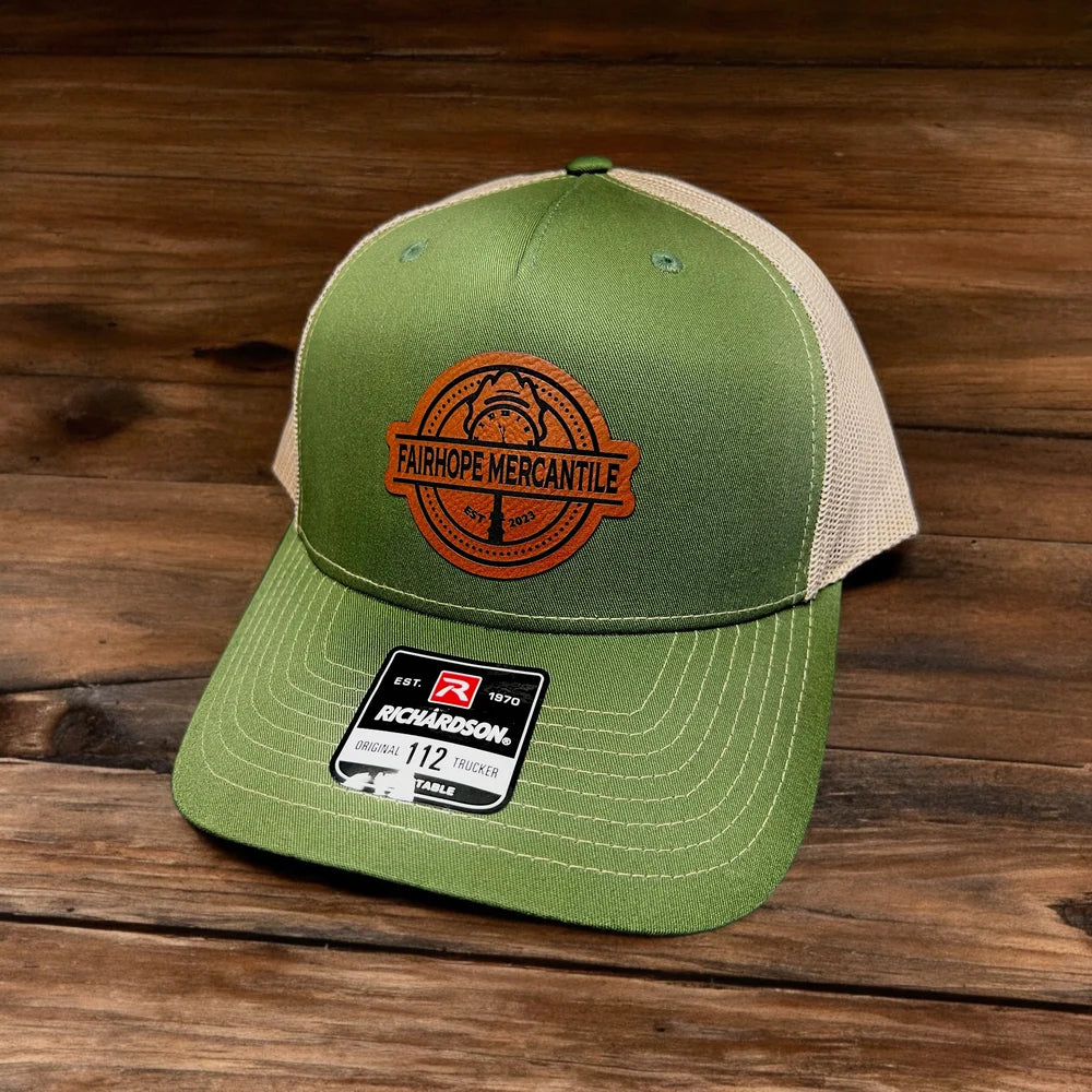 Fairhope Mercantile Five Panel Patch Hat - Richardson Trucker 112FP, featuring a green and beige color scheme with a "Fairhope Mercantile" patch on the front, placed on a wooden surface.