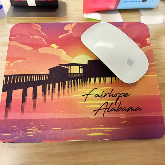 A white computer mouse rests on the Fairhope Sunset - Mousepad, which features an illustration of a sunset backdrop over Fairhope Pier, the text "Fairhope Alabama," and an anti-slip natural rubber base.