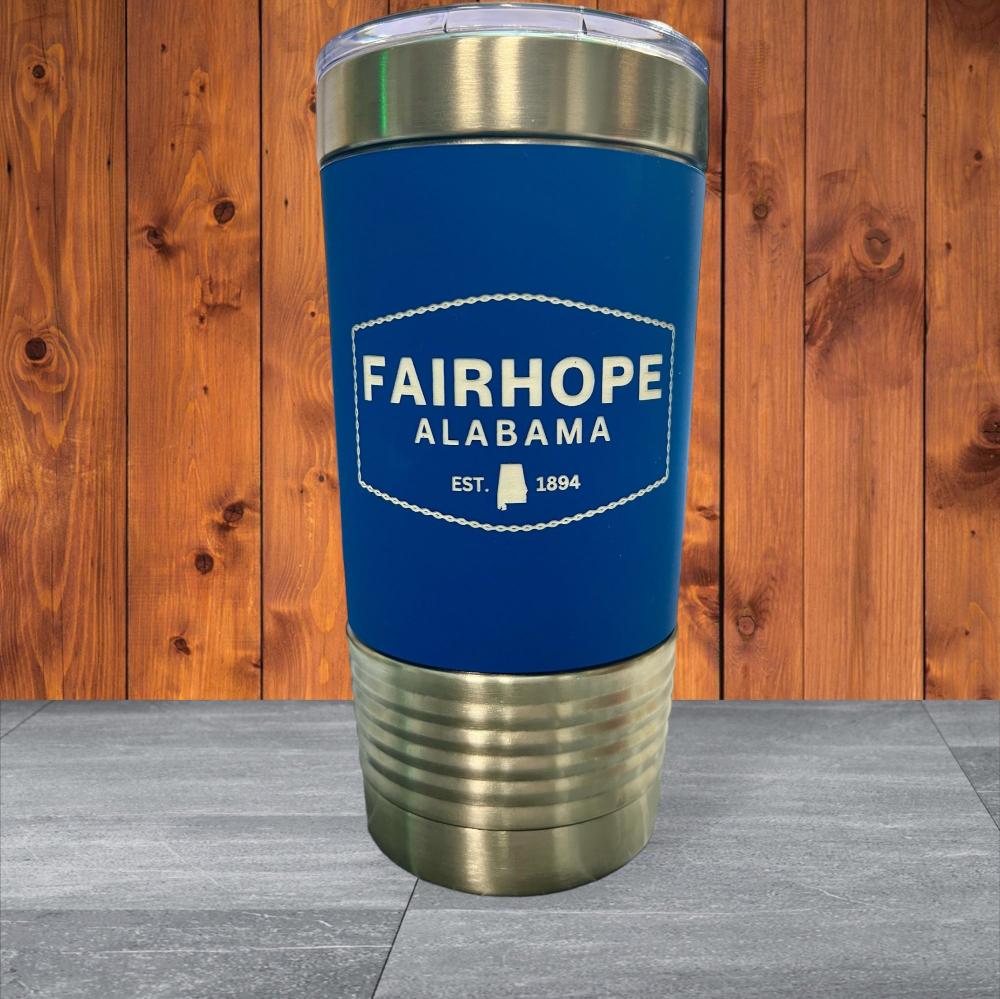 The 20oz. Stainless Tumbler with Silicone Grip features a blue exterior with a silver base and lid, adorned with the text "Fairhope Alabama Est. 1894" on a wooden background, and boasts double-wall vacuum insulation for superior temperature retention.