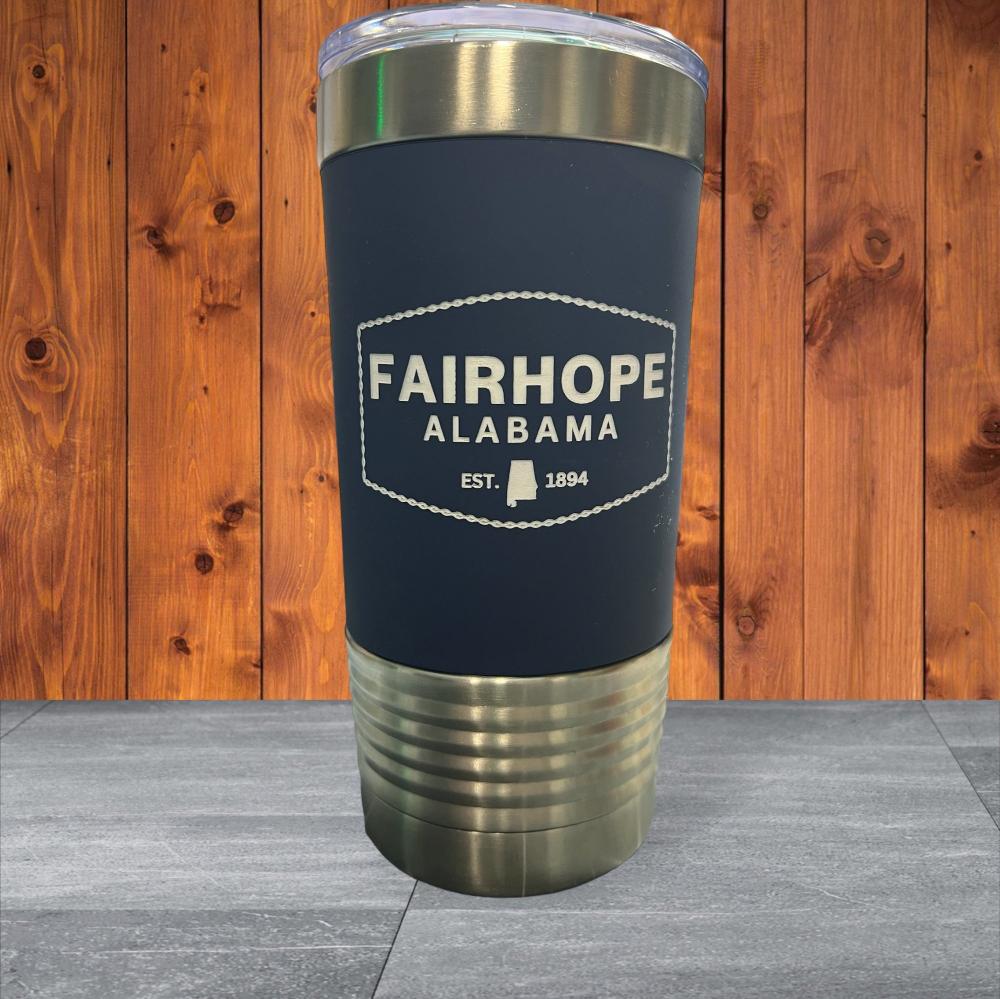 A 20oz. Stainless Tumbler with a dark blue silicone grip, adorned with "Fairhope Alabama Est. 1894" text on the front, features a silver base and lid. Crafted from stainless steel with double-wall vacuum insulation, it sits beautifully against a wooden background.