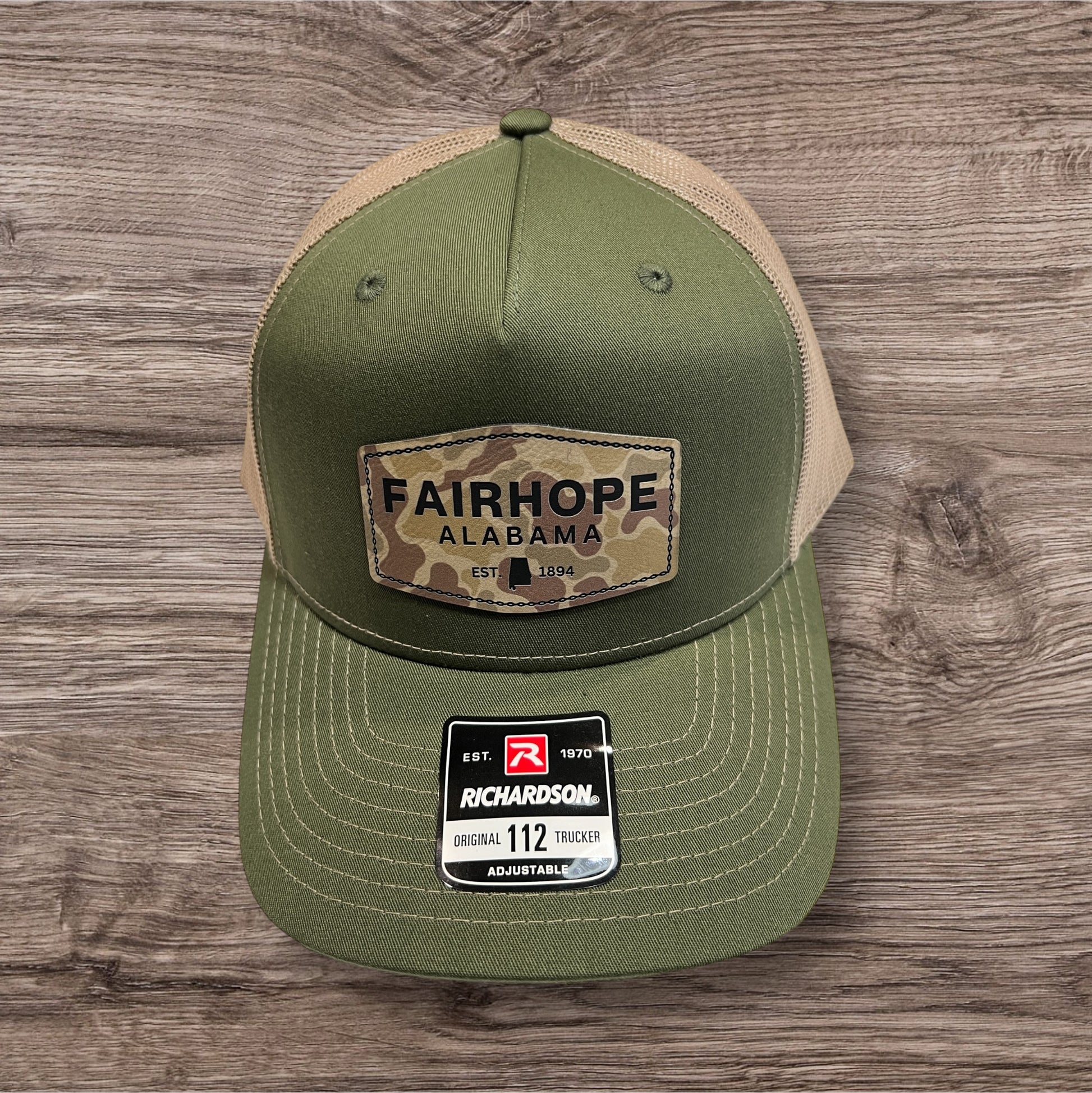 The Fairhope Heritage Logo Five Panel Patch Hat, a Richardson Trucker 112FP in green and beige, features "Fairhope Alabama Est. 1894" on a camo Fairhope Heritage patch on the front. The five-panel design hat is showcased on a wooden surface.