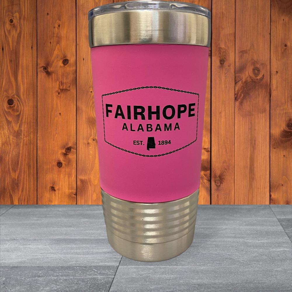 A 20oz. Stainless Tumbler with a pink Silicone Grip is adorned with a silver lid and base. Its double-wall vacuum insulation keeps beverages at the ideal temperature. The tumbler features the text "Fairhope Alabama EST. 1894" against a wooden background, adding an appealing detail to its stainless steel design.