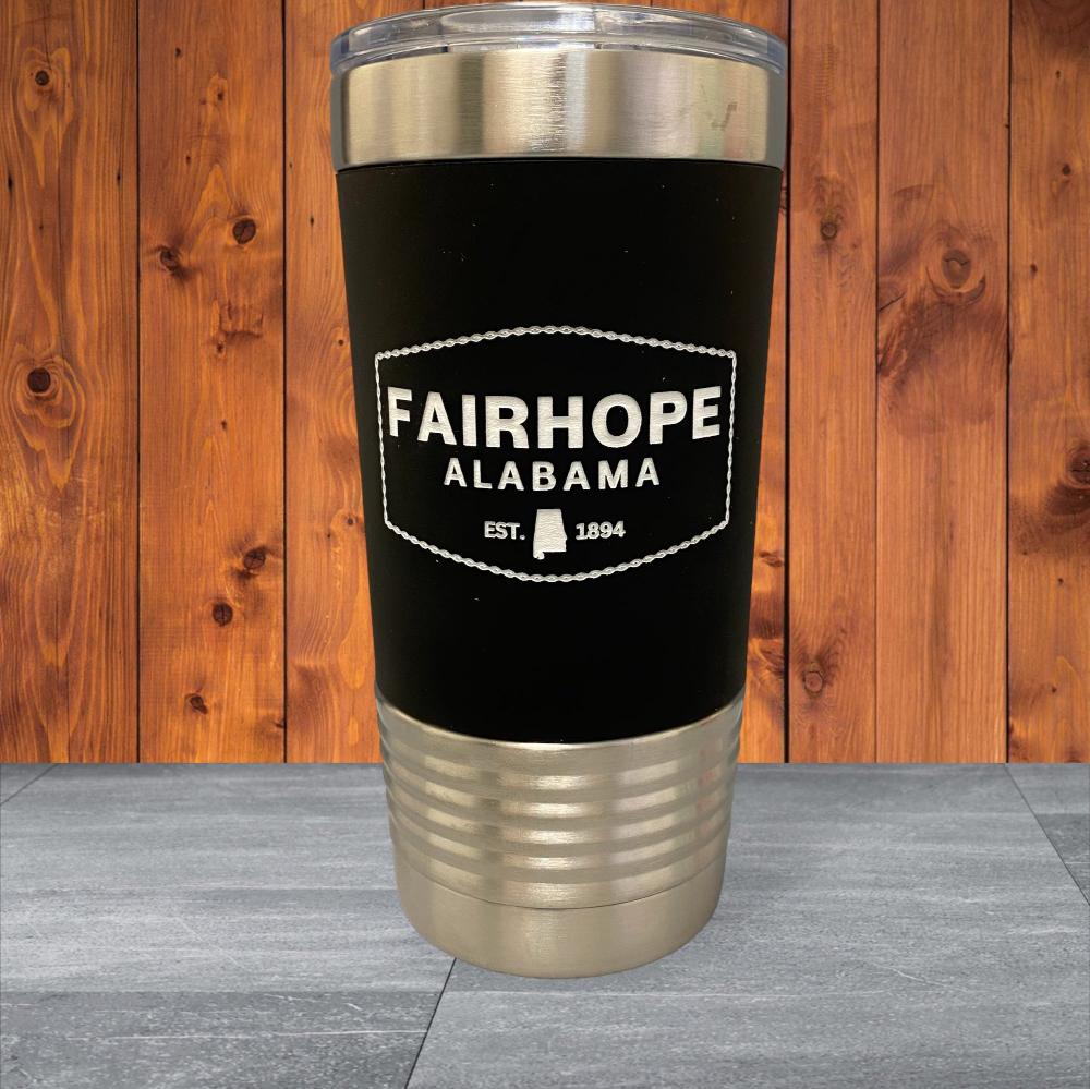 A 20oz. black and silver Stainless Tumbler with a Silicone Grip, featuring the text "Fairhope Alabama EST. 1894" in white, is set against a wooden background. The double-wall vacuum insulation ensures your drink stays at the perfect temperature.