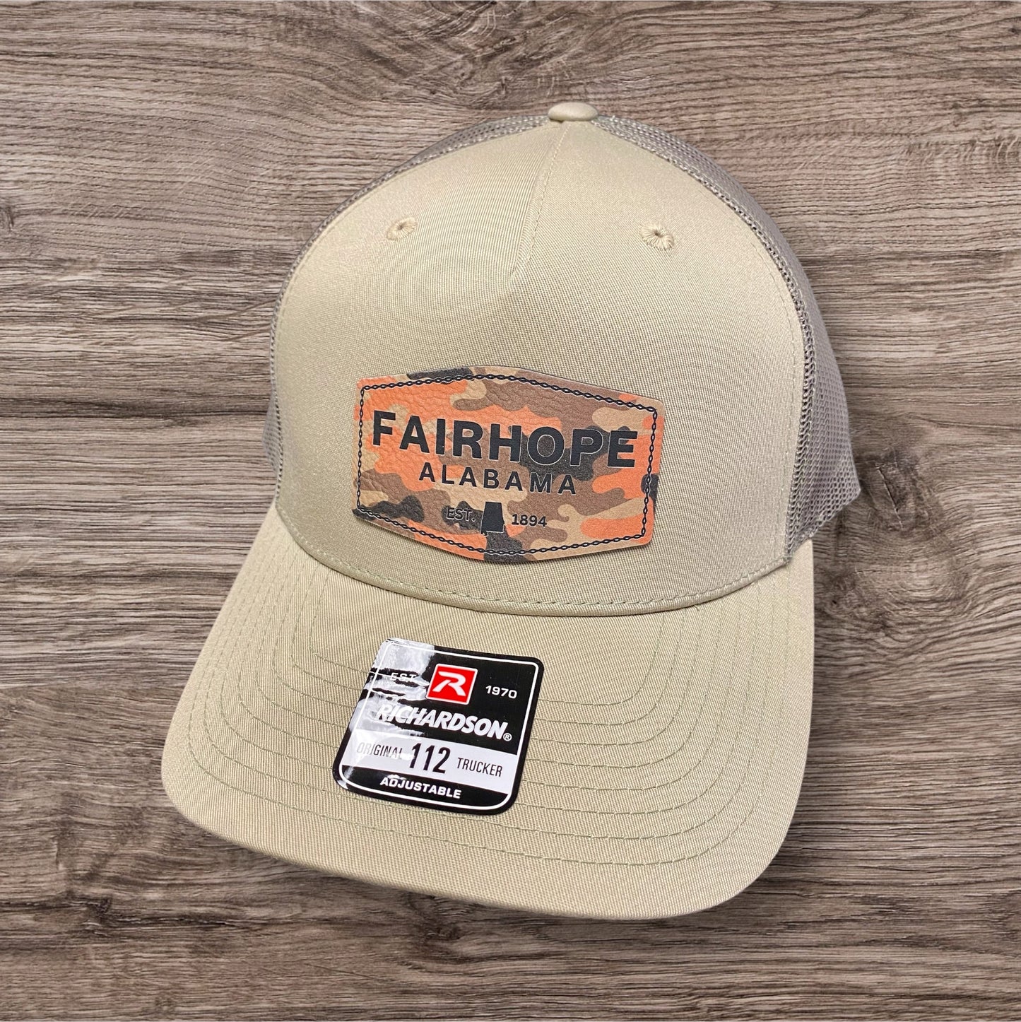 A Fairhope Heritage Logo Five Panel Patch Hat - Richardson Trucker 112FP, featuring a khaki mesh trucker hat with a camo patch reading "Fairhope Alabama 1894," is placed on a wooden surface.