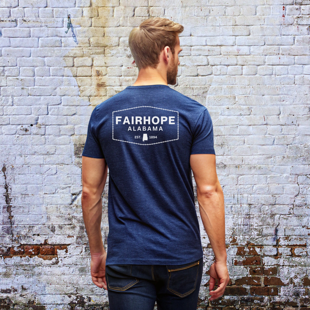 A man with short hair and a beard stands facing a brick wall, featuring the Fairhope Heritage Logo on his blue premium tee. The shirt displays "Fairhope Alabama Est. 1894" on the back and is designed for unisex sizing.