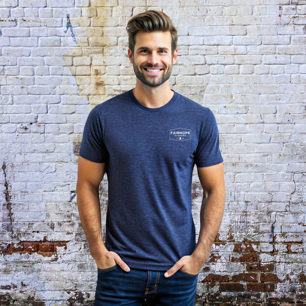 A man with brown hair and a beard is smiling while standing against a brick wall, dressed in jeans and a dark blue Fairhope Heritage Logo Tee.