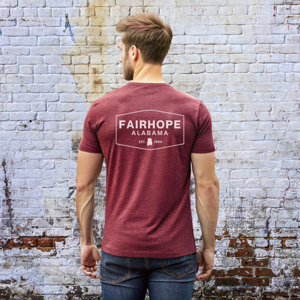 A man wearing a maroon Fairhope Heritage Logo Tee stands facing a textured brick wall.