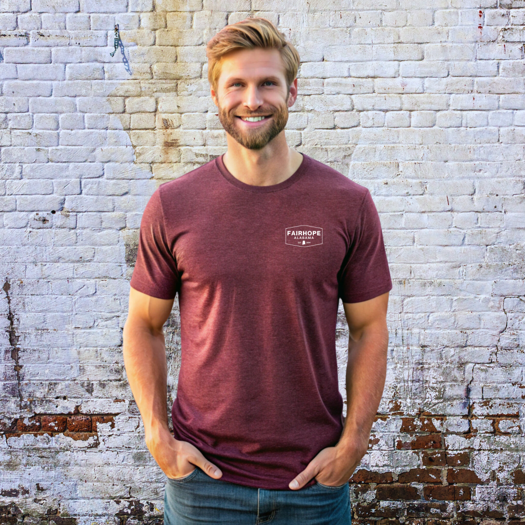 A man with a beard stands against a weathered brick wall, wearing a maroon Fairhope Heritage Logo Tee in unisex sizing and jeans, smiling with hands in pockets.