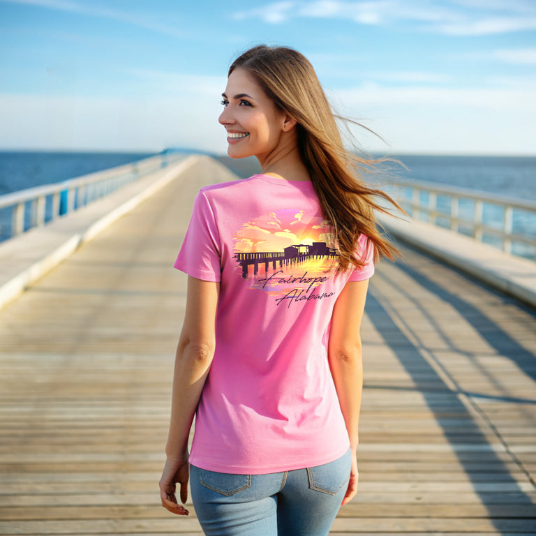 A woman in a pink Fairhope Sunset - Tee with beach imagery stands on Fairhope Pier, smiling and looking over her shoulder with Mobile Bay and the sky in the background.