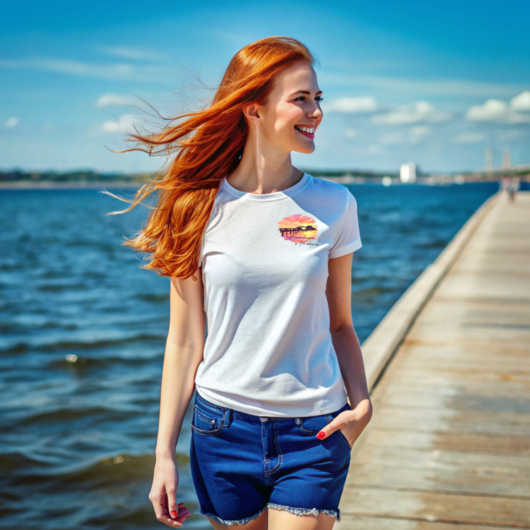 A woman with long red hair walks on Fairhope Pier by Mobile Bay, wearing a Fairhope Sunset - Tee and blue shorts. The sky is clear and sunny.