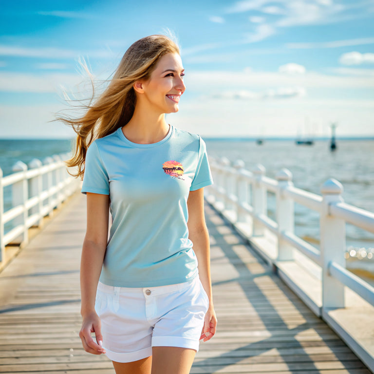 A woman in a light blue Fairhope Sunset - Tee and white shorts walks along Fairhope Pier by the sea on a sunny day.