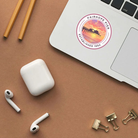 A laptop adorned with a weather-resistant vinyl "Fairhope Pier Sticker" beside AirPods, a closed charging case, gold binder clips, and two pencils on a brown surface.