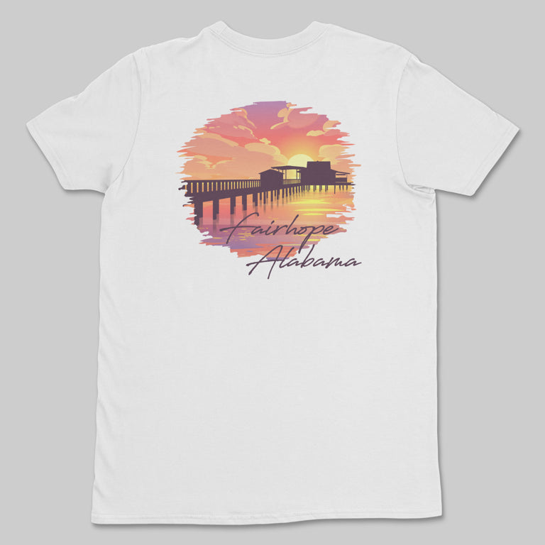 The Fairhope Sunset - Tee is a white t-shirt showcasing a sunset design with the Fairhope Pier and water reflecting the sky, accompanied by the text "Fairhope, Alabama" below the image.