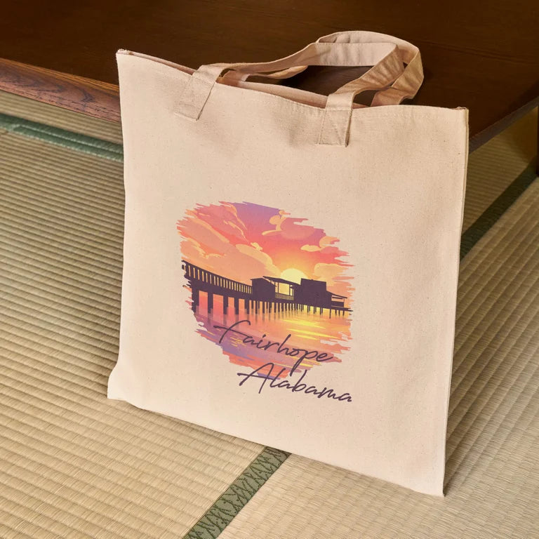 The "Fairhope Sunset - Canvas Tote," a beige canvas bag featuring an illustrated sunset and dock scene labeled "Fairhope, Alabama," rests on a woven mat, exuding coastal charm.